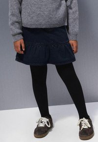 Jeanskrilo s volančki na robu, temno modre barve, v kombinaciji s črnimi legginsami in rjavimi supergami z belimi vezalkami, oblečeno s sivim puloverjem.