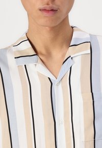 Chemise à manches courtes avec col, présentant des rayures verticales en beige, blanc et bleu clair. Comprend une poche avant et une fermeture par boutons. Tissu lisse.