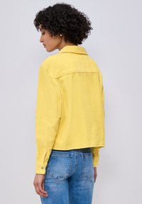 Femme aux cheveux bouclés portant une veste en velours côtelé jaune et un jean bleu, vue de dos sur un fond clair uni.