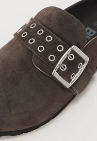 Bruine suède instap schoenen met een brede band met zilveren ogen en een grote metalen gesp. Zachte textuur en afgeronde vorm.