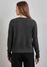 Pull en gris foncé avec des poignets et un ourlet côtelés. Texture douce, col rond et détail de couture au dos. Porté sur un pantalon noir.