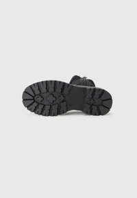 Semelle de botte noire montrant un design de crampons robuste avec des motifs distincts et une partie supérieure lisse, sur un fond blanc uni.