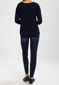 Marinblå långärmad tröja med avslappnad passform, prydd med texturerade detaljer på axlarna, i kombination med mörka jeans med vita sidostråkar och svarta skor.