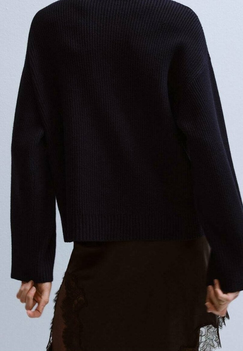 Personne portant un pull en maille noir et une jupe noire avec une bordure en dentelle, vue de dos sur un fond clair uni.