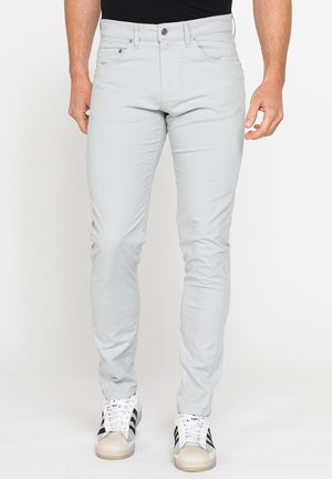 Uomo che indossa pantaloni slim fit grigio chiaro e sneakers bianche con strisce nere su uno sfondo chiaro uniforme.
