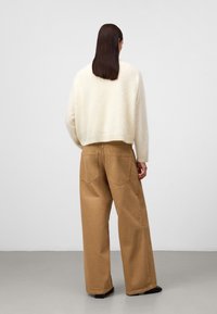 Cremfarbener, flauschiger, kürzer geschnittener Pullover; beiger, weiter Hosen mit Gesäßtaschen; glatte Textur; Modell dreht sich weg, schwarze Schuhe.
