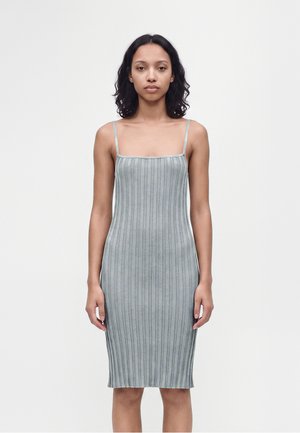 Femme aux cheveux bouclés mi-longs portant une robe grise côtelée sans manches ajustée, debout devant un fond clair uni.