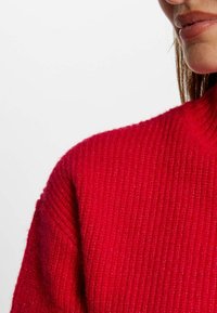 Pull en tricot rouge à col roulé avec une texture côtelée et une coupe ajustée. Le tissu semble doux avec un léger brillant.