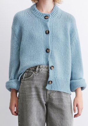 Persoon draagt een dikke lichtblauwe cardigan met knopen en opgerolde mouwen, gecombineerd met een hooggetailleerde vervaagde grijze spijkerbroek, staand tegen een effen achtergrond.