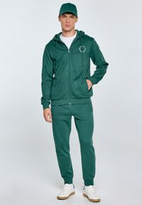 Zelená mikina na zip s dvěma kapsami, bílým logem a odpovídajícími kalhotami. Oblečení je vyrobeno z měkké tkaniny, v kombinaci s bílými teniskami.