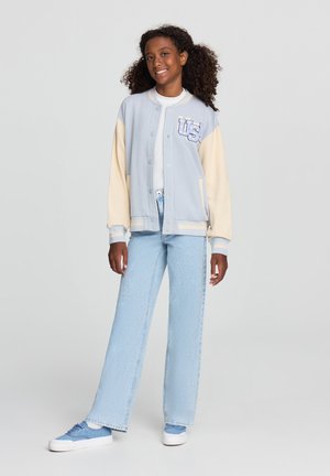 Giacca varsity blu chiaro con maniche crema, bottoni frontali e ricamo 'US'. Abbinata a jeans blu chiaro e sneaker blu.