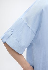 Λεπτομέρεια ανοιχτό μπλε πουκάμισου με κοντό μανίκι που δείχνει την κουμπωμένη ταινία μανικιού και το χέρι σε απλό φωτεινό φόντο.