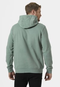 Sudadera verde menta con capucha ajustable, tela texturizada, mangas largas y puños acanalados, combinada con pantalones negros, mostrando la vista trasera.