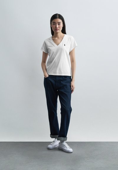 Νεαρή γυναίκα με λευκό T-shirt με V-λαιμόκοψη, σκούρο τζιν με ρεβέρ και λευκά αθλητικά παπούτσια, στεκόμενη με τα χέρια στις τσέπες μπροστά σε ένα απλό φόντο.