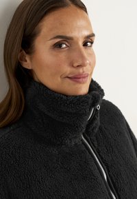 Schwarze Fleecejacke mit hohem Kragen, ausgestattet mit einem Reißverschluss, weicher Textur und einem gemütlichen Aussehen. Nahaufnahme einer Person, die sie trägt.