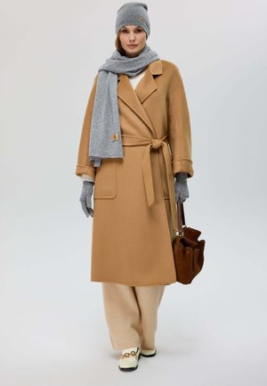 Langer beiger Mantel mit Bindegürtel, grauem Schal, passender Mütze, grauen Handschuhen, cremefarbenen Hosen und cremefarbenen Schuhen mit Goldakzent. Braune Tasche.