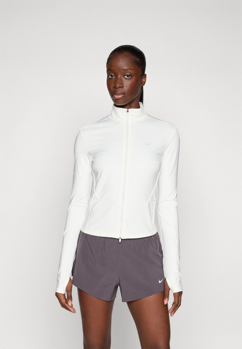 Hvit zip-up treningsjakke med lange ermer og høy krage, kombinert med mørkegrå shorts som har en liten logo. Glatt, tettsittende stoff.