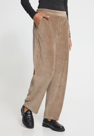 Frau trägt beige Cord-Hose mit weitem Bein, schwarzes Langarmoberteil und schwarze Leder-Loafer, steht mit einer Hand in der Tasche.