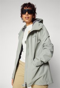 Veste imperméable gris clair avec capuche, dotée d'une fermeture éclair, de poches latérales et d'une doublure intérieure contrastante. Portée par-dessus une chemise blanche.