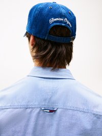 Denimblauwe pet met "American Original" in het wit geborduurd, achterstevoren gedragen, boven een lichtblauw shirt met gekleurde accentdetails.