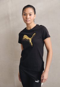 Tricou din bumbac negru cu un design al logo-ului Puma aurie. Modelul îl asortează cu colanți negri, completând un look athletic simplu.