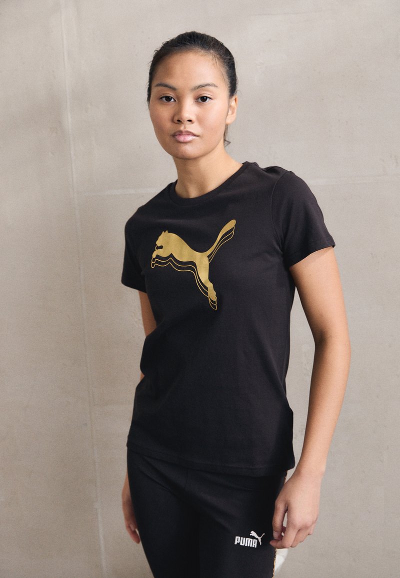Tricou din bumbac negru cu un design al logo-ului Puma aurie. Modelul îl asortează cu colanți negri, completând un look athletic simplu.