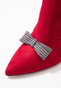 Chaussure pointue en suède rouge avec un accent en nœud façon houndstooth noir et blanc. Caractéristiques : texture lisse et couture soignée.