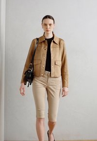 Suedes brun jakke med knapp foran og lommeflater, kombineres med beige tettsittende caprisbukser, svart topp og en liten svart håndveske.