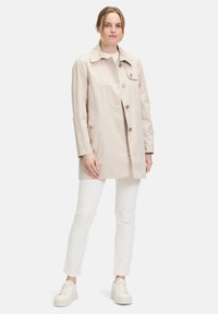 Beige wasserdichter Mantel mit Kragen, vorderen Knöpfen, zwei Seitentaschen und leicht A-Linien-Schnitt, kombiniert mit weißen Hosen und Sneakers.