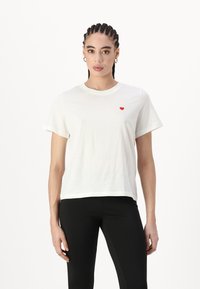 Witte katoenen T-shirt met korte mouwen, voorzien van een klein rood hartlogo op de linkerkant van de borst. Rechte snit, zachte textuur en een minimalistisch ontwerp.