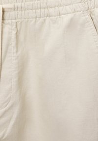 Shorts en tissu beige clair avec taille élastique et poche latérale, montrant les détails de la texture et des coutures.