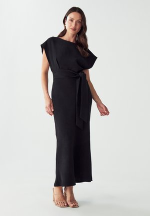 Black maxi dress with short, wide sleeves, a belted waist, and a straight silhouette. Smooth texture; mid-length side slits. Nude high-heeled sandals.
