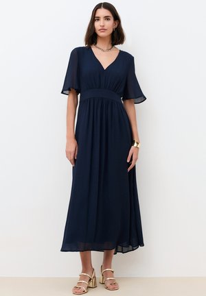 Femme portant une robe midi bleu marine à encolure en V avec manches courtes flottantes, collier chaîne en or, bracelet manchette, et sandales à talons dorés.