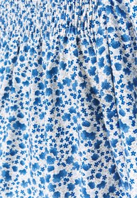Blåt og hvidt blomstret stof med smockdetaljer, med forskellige nuancer af blå blomster på en hvid baggrund.