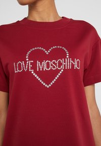 Personne portant un T-shirt rouge avec le texte "LOVE MOSCHINO" et un contour de cœur en clous argentés sur la poitrine.
