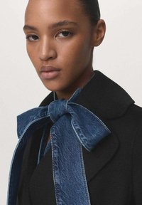 Manteau noir avec un grand nœud papillon en denim. Le nœud a une finition en denim texturé et des coutures visibles. Visage du mannequin visible, fond neutre.