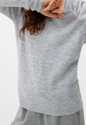 Person iført en blød grå sweater med ribstrikkede manchetter og kant, der delvist dækker deres ansigt med sammenflettede hænder.