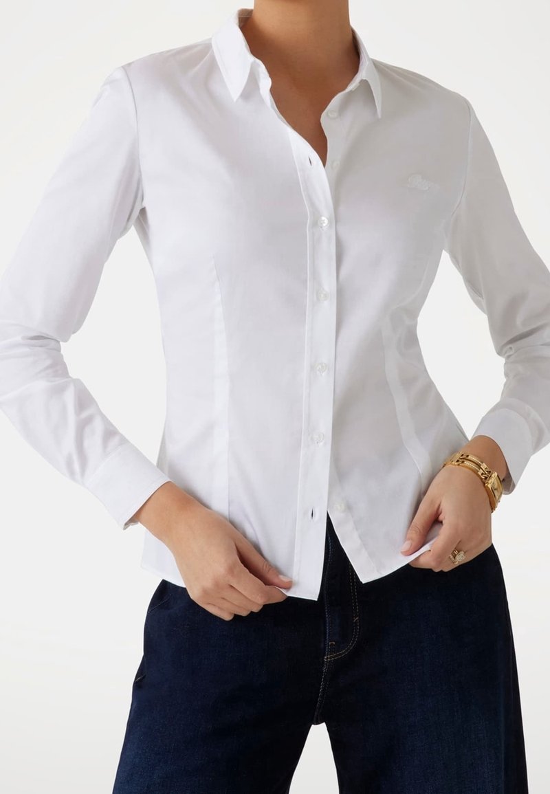 Personne portant une chemise blanche cintrée à boutons et un pantalon bleu foncé, avec des bracelets en or et une bague à la main gauche.