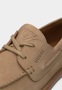 Mocassin en daim beige avec détails cousus, bout rond, devant à lacets, logo GUESS en relief sur le côté, et patch triangulaire de la marque sur la languette.