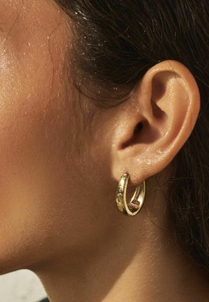 Nahaufnahme vom rechten Ohr einer Person mit einem strukturierten goldenen Creolen-Ohrring, glatter Haut und zurückgebundenem dunklem Haar.