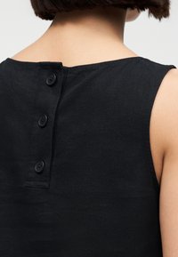 Πίσω όψη ενός ατόμου με κοντά καστανά μαλλιά που φοράει ένα μαύρο αμάνικο τοπ με τρία μαύρα κουμπιά στον άνω μέρος της πλάτης.