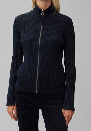 Frau trägt einen taillierten, gerippten, marineblauen Reißverschluss-Pullover mit langen Ärmeln und hohem Kragen vor einem schlichten grauen Hintergrund.