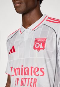 Homme portant un maillot de football Olympique Lyonnais blanc avec des logos rouges, un texte sponsor Emirates rose et un col boutonné.