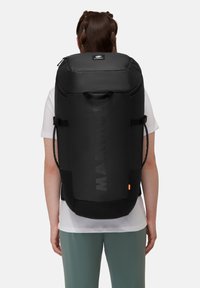Czarny plecak z wyściełanego materiału, z regulowanymi paskami, uchwytem u góry i wyróżniającym się logo z przodu. Smukły, cylindryczny kształt z minimalnymi akcentami.