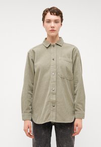 Chemise en velours côtelé vert avec col, manches longues et poche avant. Caractéristiques : boutons en métal argenté et tissu texturé.