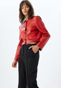 Giacca in pelle rossa corta con scollatura tonda, chiusura frontale con bottoni e una tasca, abbinata a pantaloni neri a vita alta.
