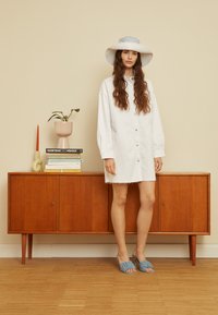 Vestido de camisa em algodão branco com frente em botões e bainha desfiada. Sapatos azul claro de calçar. Fundo neutro com armário de madeira e plantas.
