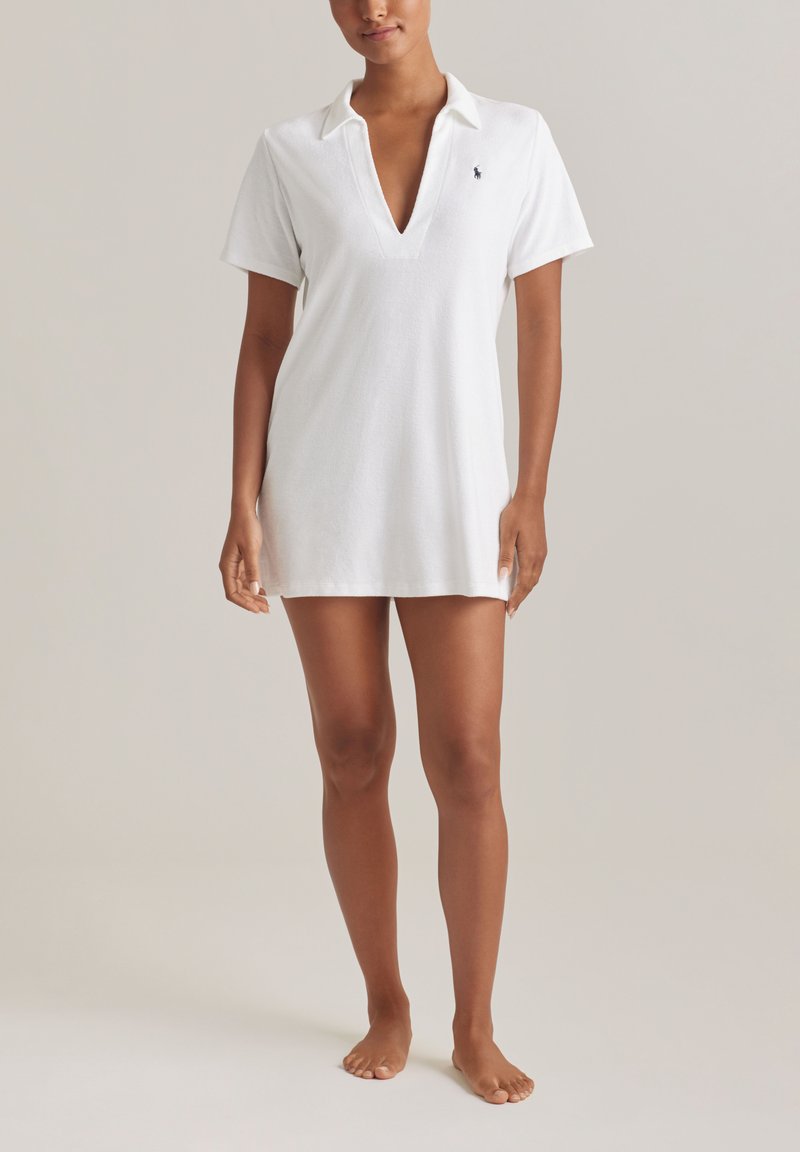 Γυναίκα που φοράει ένα λευκό φόρεμα από περκάλι με κοντά μανίκια, γιακά, βαθύ V στο ντεκολτέ και μικρό κεντημένο λογότυπο στο στήθος, όρθια, ξυπόλητη.