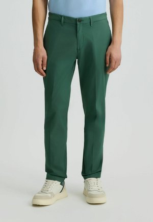 Green trousers made of smooth fabric, featuring a straight cut, front pockets, and a brown button closure. Paired with beige sneakers.
