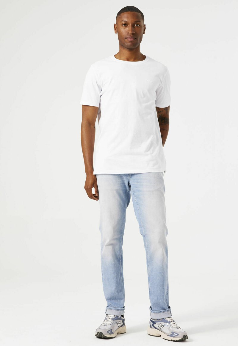 Camiseta blanca de manga corta combinada con jeans de cortos azules. La camiseta tiene un diseño de cuello redondo. El modelo lleva zapatillas grises con acentos oscuros.